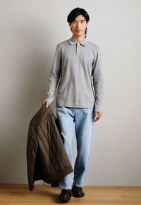 Polo shirt grigio a maniche lunghe con colletto, jeans azzurri, e giacca marrone trapuntata tenuta in mano. Scarpe scure, sfondo neutro.