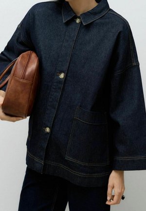 Person wearing dark denim jacket with large front pocket, holding brown leather bag under left arm against plain background.