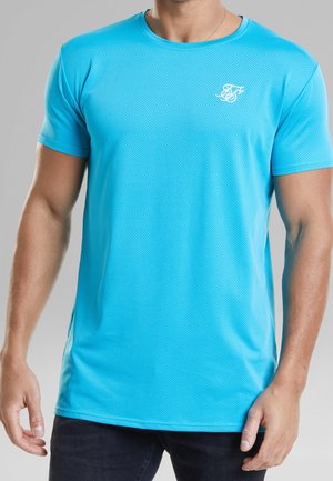 Mężczyzna ubrany w jasnoniebieski teksturowany t-shirt z krótkim rękawem i małym białym logo na lewej piersi, ciemne spodnie i srebrny naszyjnik.