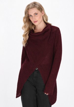 Donna bionda con capelli mossi indossa un maglione a maglia asimmetrico bordeaux sopra pantaloni neri, mentre posa su uno sfondo bianco.
