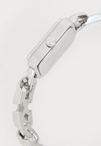 Ασημένιο μεταλλικό ρολόι χειρός, πλευρική όψη που δείχνει την κορώνα με χαραγμένο λογότυπο και μπρασελέ με σχέδιο κοπής βέλους.