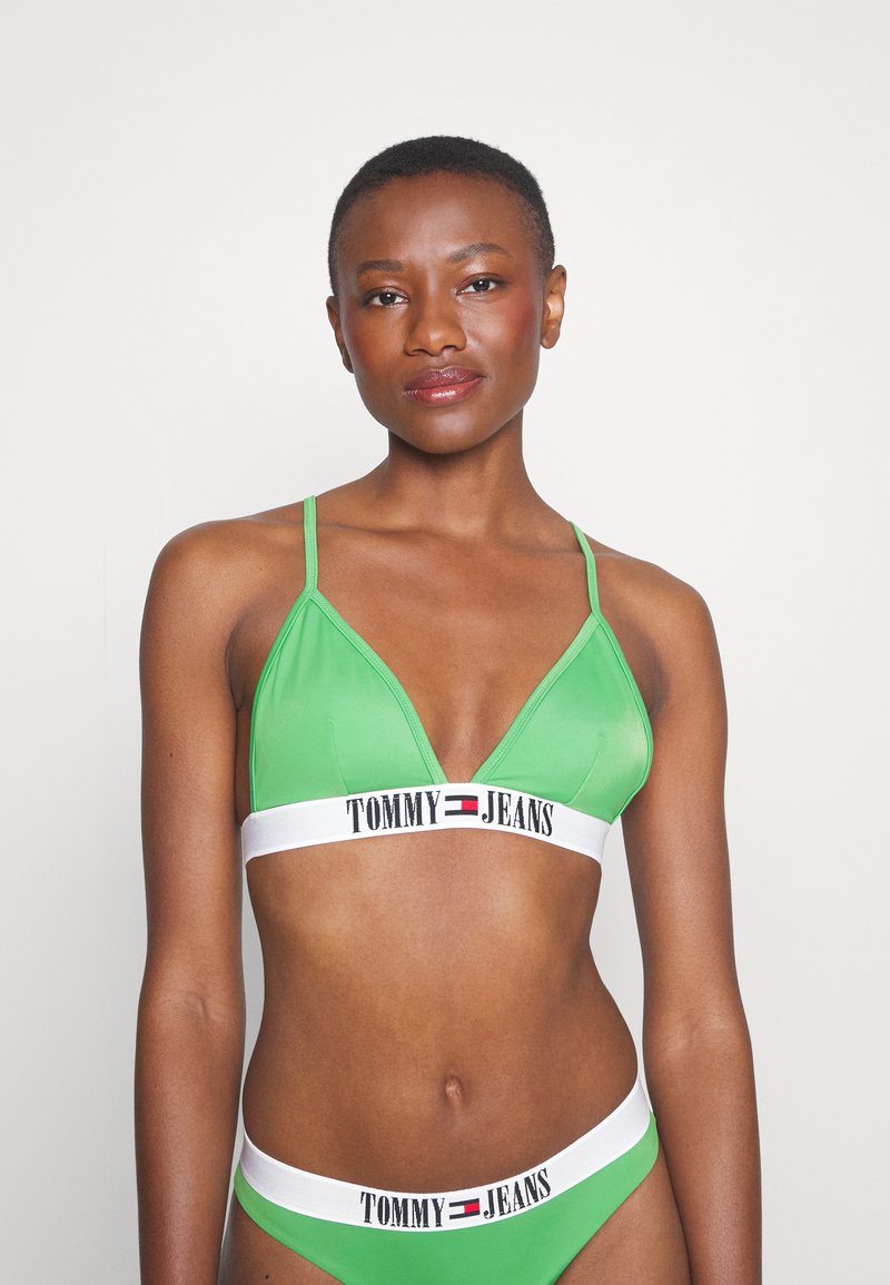 Zelený trojuholníkový bralette s tenkými ramienkami, ktorý má biely elastický pás s potlačou "TOMMY JEANS" a logom. Hladká textúra.