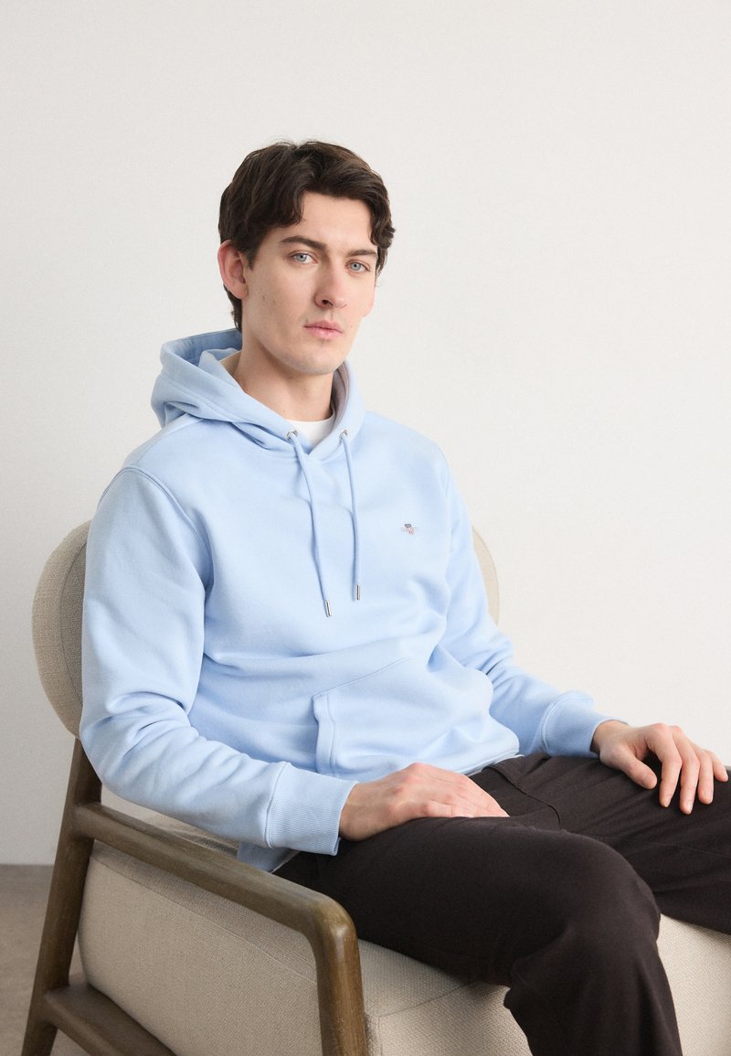 Light blue cotton hoodie with a front pocket, drawstring neck, and ribbed cuffs, paired with dark pants, seated on a wooden chair.