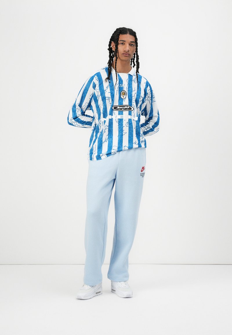 Blaue und weiße gestreifte Sweatshirt mit abstrakten Mustern, kombiniert mit hellblauen Jogginghosen mit einem Logo. Weiße Sneakers vervollständigen das Outfit.