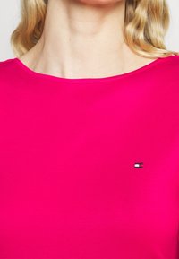Tætsiddende pink kortærmet top lavet af et glat materiale, med et lille logo i nederste venstre hjørne. Rund halsudskæring.