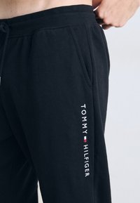Schwarze Baumwoll-Sweatpants mit einem Kordelzug in der Taille, ausgestattet mit Seitentaschen und einem weißen gestickten "TOMMY HILFIGER"-Logo entlang des Beins.