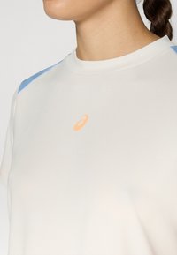 Lengvas, trumpomis rankovėmis sportinis marškinėliai kreminės spalvos su mėlynomis petų akcentais ir oranžine logotipu ant krūtinės; lygus audinio tekstūra.