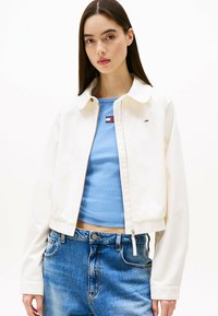Korte witte jas met een kraag, gecombineerd met een lichtblauw ribbeltop en hooggeknoopte blauwe jeans. Eenvoudig ontwerp en strakke lijnen.