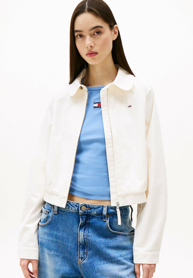 Korte witte jas met een kraag, gecombineerd met een lichtblauw ribbeltop en hooggeknoopte blauwe jeans. Eenvoudig ontwerp en strakke lijnen.