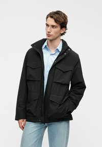 Nuori mies, joka käyttää mustaa takkia, jossa on neljä suurta etutaskua, vaaleansinisen paidan ja vaaleansinisten farkkujen päällä, katsoo oikealle käsi taskussa.