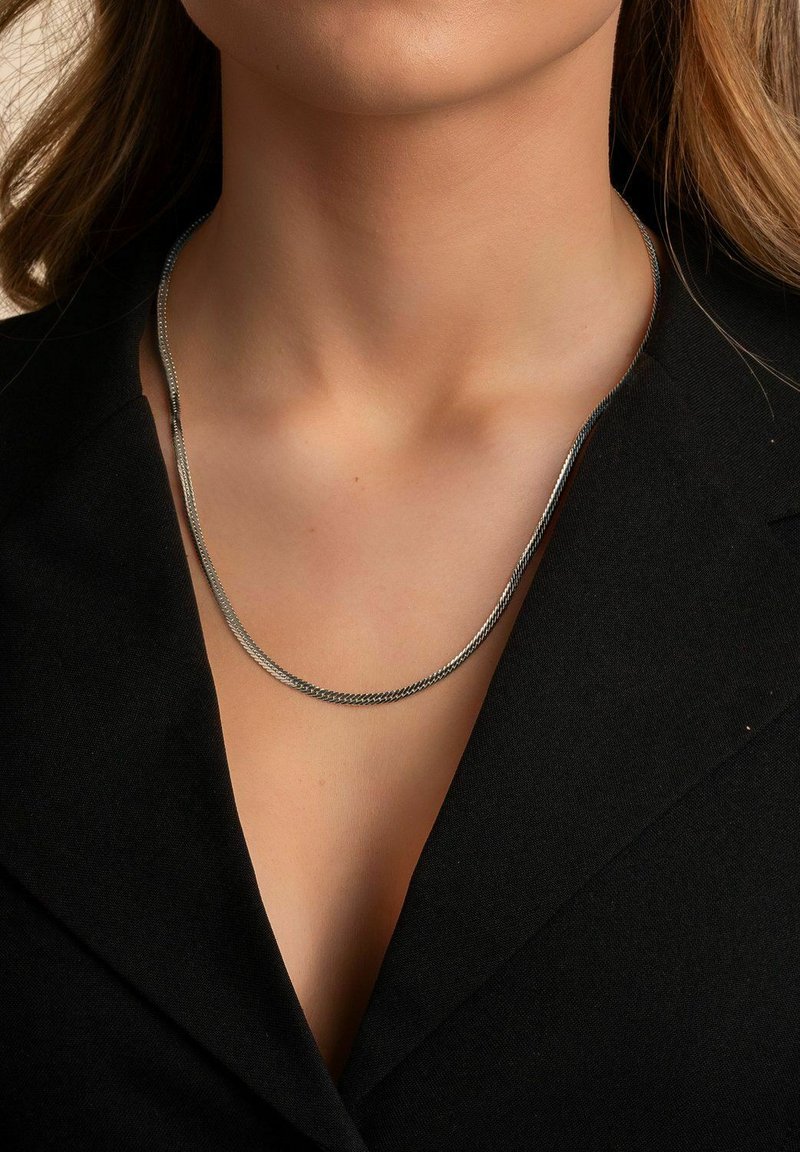 Silverfärgad kedjanecklace med två strängar, med en slät och vriden textur, vilande på ett naket nyckelben, mot en svart topp.