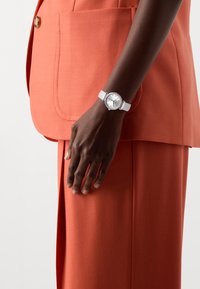Montre avec un boîtier en ton argent et un bracelet blanc, dotée d'un cadran rond avec des marqueurs et des accents en gemmes. Portée sur une main à la peau foncée contre un costume orange.