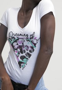 La camiseta blanca de cuello en V presenta un estampado gráfico con el texto "Soñando con GUESS", un patrón de leopardo y acentos geométricos en rosa y verde.