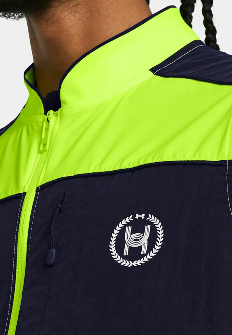 Hombre vistiendo una chaqueta con cremallera en azul marino y verde brillante, con un emblema circular blanco que presenta una "H" y una corona de laurel en el pecho.