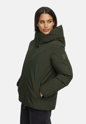 Vrouw met donker haar die een donkergroene gewatteerde jas met hoge kraag draagt, handen in de zakken, kijkend naar de zijkant tegen een effen achtergrond.