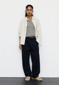 Młoda kobieta stoi przy białej ścianie, ubrana w beżową, pasiastą, rozpiętą koszulę, szary krótki sweter, luźne czarne spodnie i beżowe trampki.