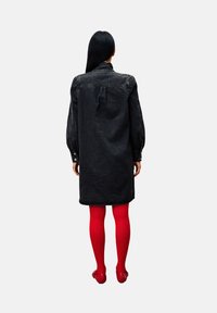 Vrouw met de rug naar de kijker, gekleed in een zwarte denim jurk, felrode panty en rode schoenen, tegen een effen lichte achtergrond.