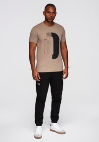 Vaaleanbeige T-paita, jossa on musta graafinen design, sisältäen tekstiä ja muotoja, yhdistettynä mustiin cargo-housuihin ja valkoisiin lenkkikengiin, joissa on harmaita yksityiskohtia.