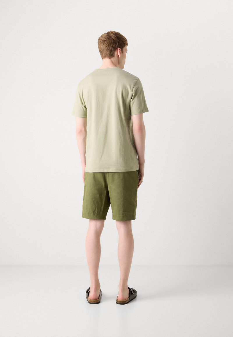 T-shirt en coton vert olive et shorts assortis. Le T-shirt a un col rond ; les shorts ont une coupe décontractée et un minimum de détails. Sandales noires.
