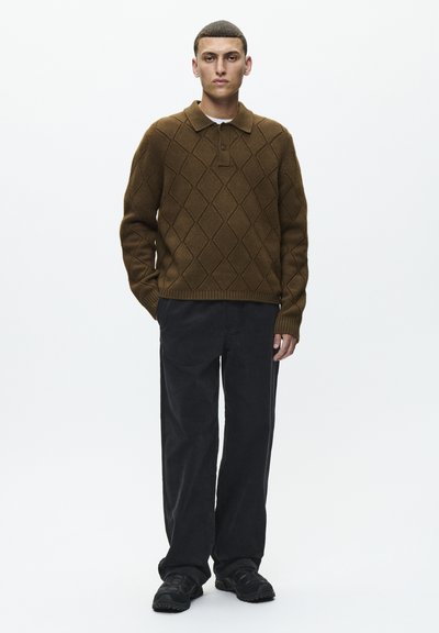 Pull en polo tricoté brun avec un motif en losanges, associé à un pantalon en velours côtelé noir et des baskets noires, présenté sur un fond blanc.