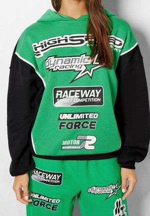 Vrouw die een groene en zwarte hoodie en broek draagt met racethema-tekst en -grafieken, waaronder "Raceway," "Unlimited Force," en "Dynamic Racing."