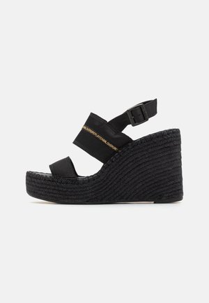 Des sandales à talons noirs présentent une plateforme tissée, des sangles élastiques et une boucle sur le côté. Le design comprend une finition lisse et un minimalisme dans les détails.
