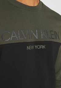 Sudadera de cuello redondo con una sección superior de color verde oliva y una sección inferior negra. Presenta un texto en negrita, gris oscuro, que dice "CALVIN KLEIN" y "NEW YORK".