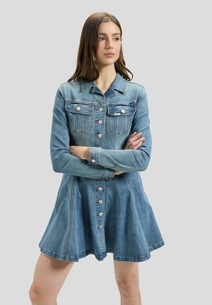 Junge Frau mit langen braunen Haaren trägt ein hellblaues Denimkleid mit langen Ärmeln und Knöpfen, steht mit verschränkten Armen und blickt nach rechts.