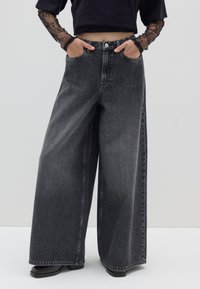 Pantalones anchos de mezclilla negra con cintura alta, que presentan bolsillos delanteros y un sutil desgaste en la tela. Combinados con un top negro corto.