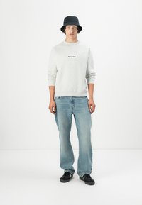 Lichtgrijze sweatshirt met geborduurde tekst, blauwe jeans, zwarte bucket hat en zwarte sneakers met witte accenten; relaxte pasvorm.