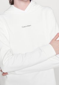 Sudadera blanca con capucha hecha de tela suave, con un pequeño logo "Calvin Klein" en gris oscuro en el pecho. Mangas largas con puños acanalados.