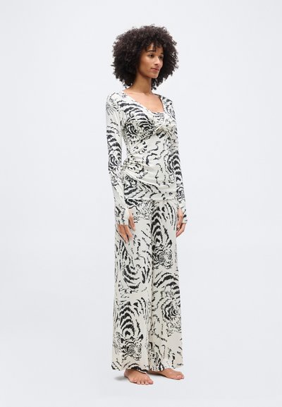 Γυναίκα με σγουρά μαλλιά που φοράει λευκό τοπ με μακριά μανίκια και παντελόνι φαρδιάς γραμμής με μαύρο σχέδιο τίγρης, όντας ξυπόλυτη μπροστά σε λευκό φόντο.