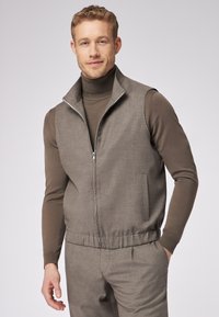 Graue ärmellose Weste mit hohem Stehkragen, Reißverschluss, strukturiertem Stoff und elastischem Bund. Wird über einem braunen Rollkragenpullover getragen.