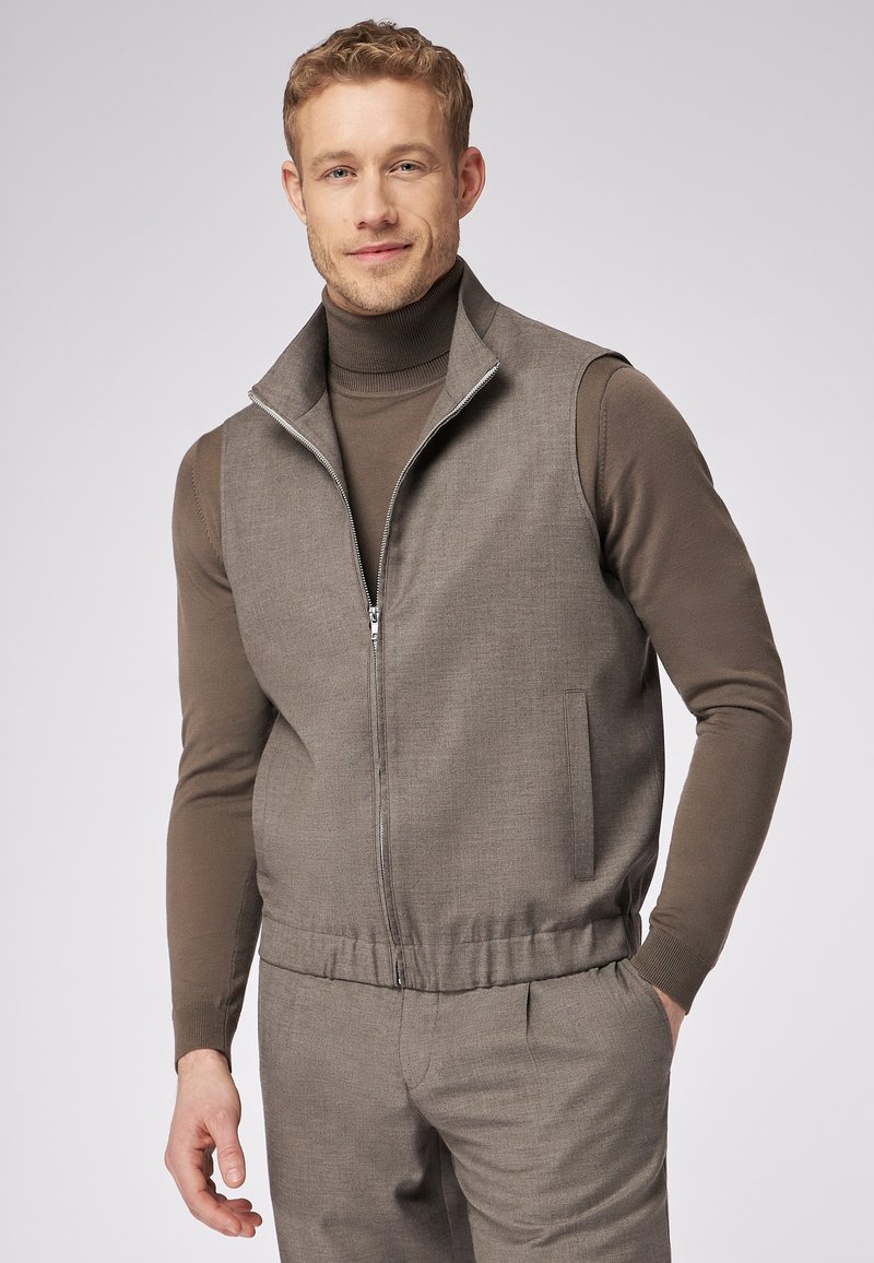 Graue ärmellose Weste mit hohem Stehkragen, Reißverschluss, strukturiertem Stoff und elastischem Bund. Wird über einem braunen Rollkragenpullover getragen.