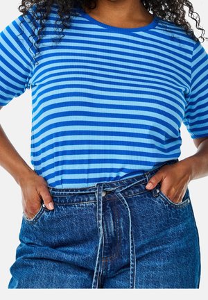 Kobieta ubrana w niebieską koszulę w paski z krótkim rękawem, wpuszczoną w wysokie niebieskie dżinsy, z rękami w kieszeniach.
