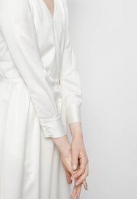 Personne portant une robe longue en satin blanc à manches longues avec des poignets boutonnés, les mains doucement jointes devant, sur un fond blanc uni.