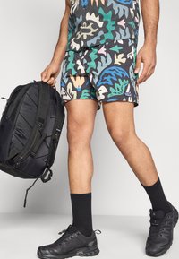 Πολύχρωμα σορτς με έντονα σχέδια φύλλων, σε συνδυασμό με μαύρο σακίδιο πλάτης, μαύρες κάλτσες και σκούρα αθλητικά παπούτσια σε ουδέτερο περιβάλλον.