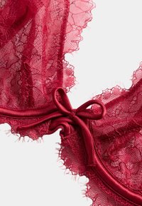 Czerwony koronkowy biustonosz z misternymi kwiatowymi wzorami, delikatnym falowanym brzegiem i satynową kokardką na środku. Miękka faktura, konstrukcja z fiszbinami.