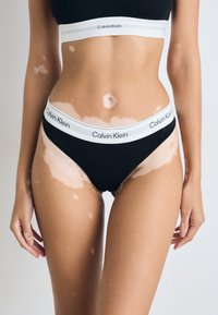 Crne pamučne bikini gaćice s bijelim Calvin Klein logotipom. Prikazane su značajne varijacije u tonovima kože na trbuhu i bedrima.