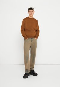 Hnedý sveter s okrúhlym výstrihom, v kombinácii so svetlými hnedými nohavicami. Čierne šnurovacie topánky a šedá čiapka dotvárajú outfit. Jednoduché pozadie.