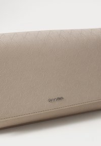 Pochette beige gaufrée avec un motif géométrique, texture lisse et un logo métallique Calvin Klein à l'avant.