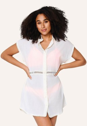 Vrouw met krullend haar staat met handen in de zij, gekleed in een witte jurk met korte mouwen, knopen en kanten rand en kraag.