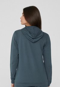 Jeune femme aux longs cheveux bruns portant un sweat à capuche bleu foncé, tournée vers la gauche, vue de dos sur un fond clair uni.