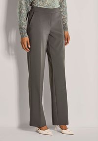 Vrouw in hoge taille, wijde grijze broek, met een gedrukt lang mouwtop en witte puntige hakken, staande tegen een effen achtergrond.