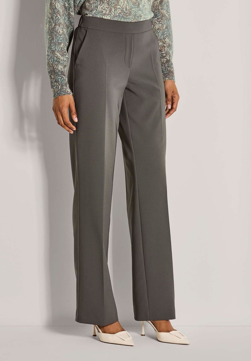 Vrouw in hoge taille, wijde grijze broek, met een gedrukt lang mouwtop en witte puntige hakken, staande tegen een effen achtergrond.