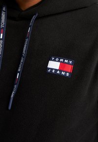 Felpa nera in pile con patch del logo Tommy Jeans, dettagli in rosso, bianco e blu, e cordini blu navy con testo del marchio.