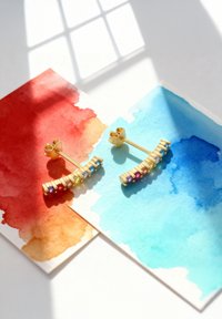 Goldene Ohrringe mit geschwungenem Design, verziert mit mehrfarbigen Edelsteinen. Auf Aquarellpapier in Rot- und Blautönen präsentiert.