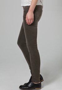 Jeans skinny verde oliva con una textura suave, cintura media y diseño clásico de cinco bolsillos, combinados con zapatos negros de cordones.