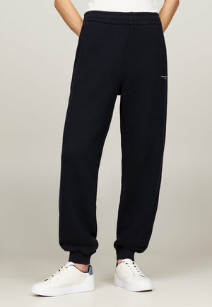 Persoon draagt zwarte joggerbroek en witte sneakers, staat met handen in de zakken tegen een effen achtergrond.