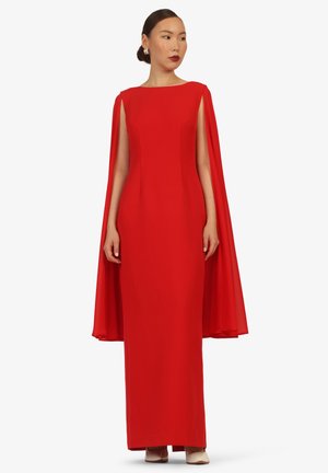Frau trägt ein bodenlanges rotes Kleid mit angenähten, fließenden Umhangärmeln, steht mit locker an den Seiten hängenden Händen und blickt zur Seite.